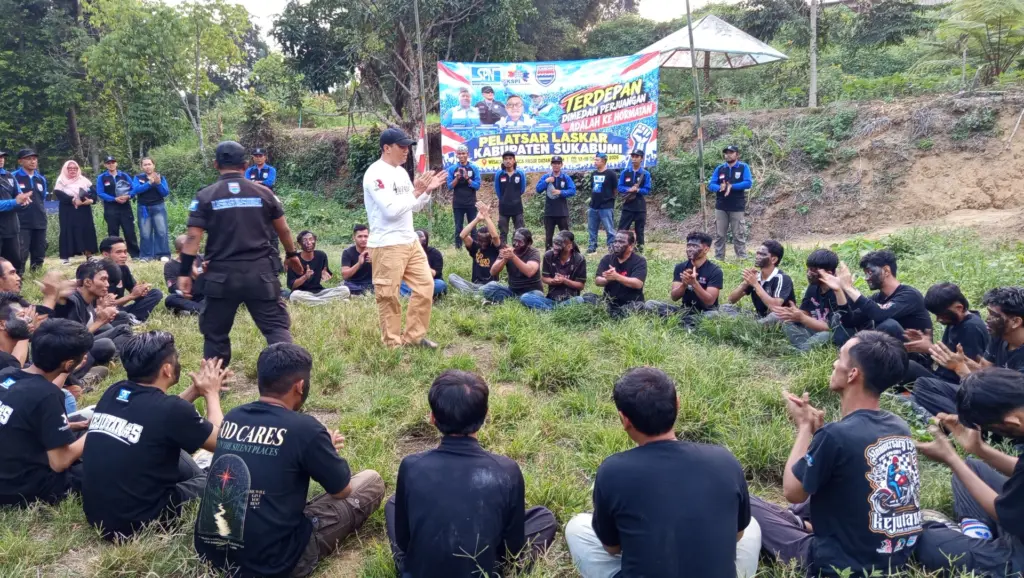 Pemateri berbicara di tengah peserta yang duduk di rumput dengan wajah dicoreng
