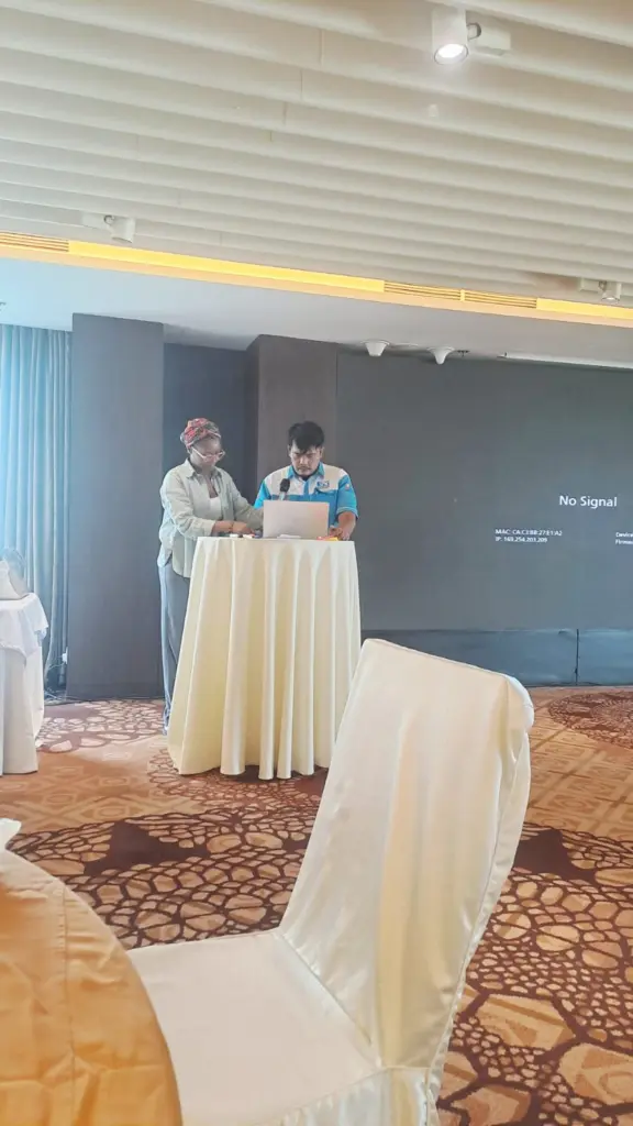Rizky dan seorang fasilitator menyiapkan laptop di meja presentasi