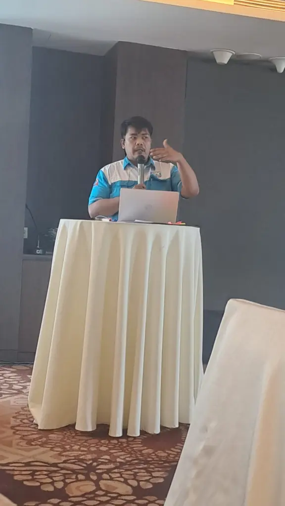 Rizky berbicara sambil memberi isyarat tangan di balik meja presentasi